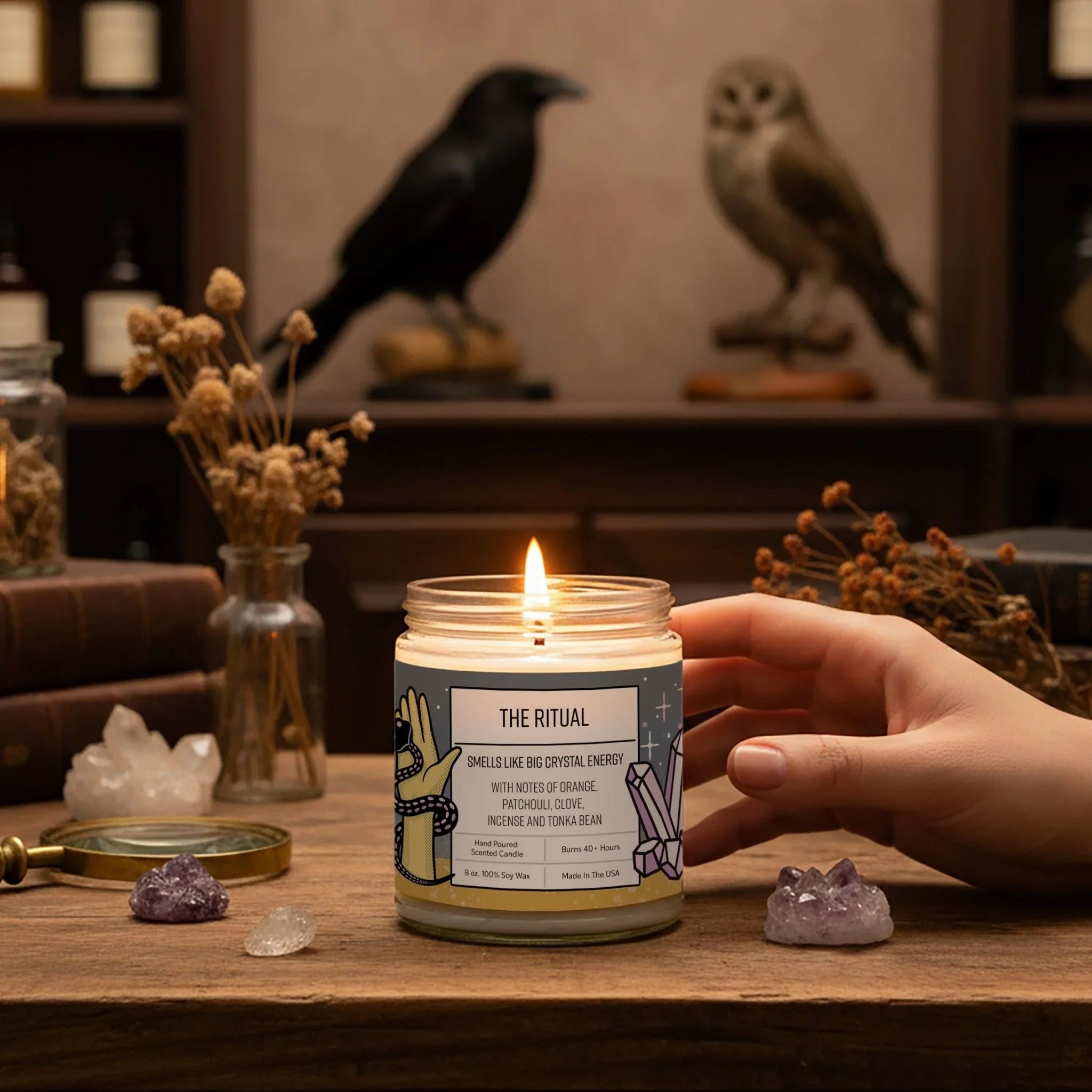 A hand is holding a lit candle labeled "THE RITUAL" on a wooden surface, surrounded by various crystals, dried flowers, and a bird statue.