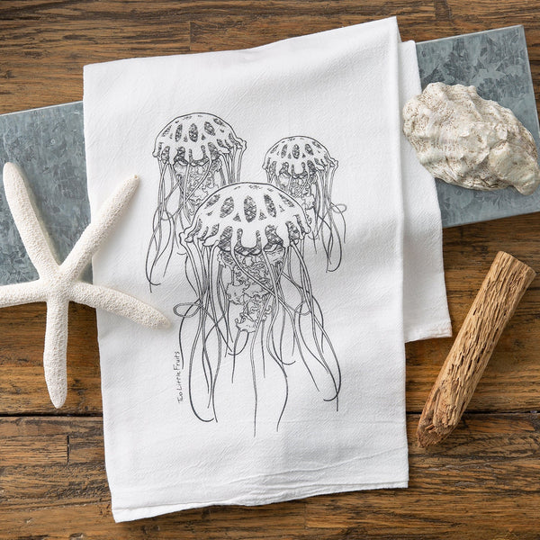A jellyfish tea towel positioned on a wooden table, featuring a charming illustration of three jellyfish, alongside a starfish and natural elements.