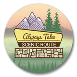 a sign that says "Always Take the Scenic Route" with mountains, pine trees, and a sunset in the background.