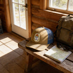 A tan baseball cap with a blue brim and a patch on the front, along with a pair of sunglasses and a map, are placed on a wooden bench in a rustic setting.