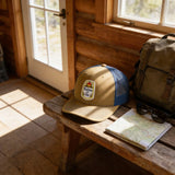 A tan baseball cap with a blue brim and a patch on the front, along with a pair of sunglasses and a map, are placed on a wooden bench in a rustic setting.