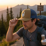 A man wearing a tan baseball cap with a mountain range in the background, adjusting his cap while carrying a backpack.