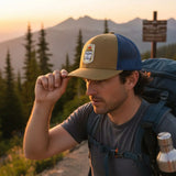 A man wearing a tan baseball cap with a mountain range in the background, adjusting his cap while carrying a backpack.