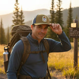 A man wearing a hat and backpack stands in a grassy field, with a signpost visible in the background.