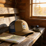 A tan baseball cap with a Nature and Shitty patch sits on a wooden bench, accompanied by a map, sunglasses, and a rope.