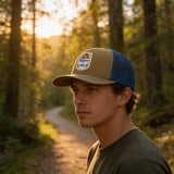 A man wearing a baseball cap with a patch stands on a dirt path in a forest, surrounded by trees and sunlight.