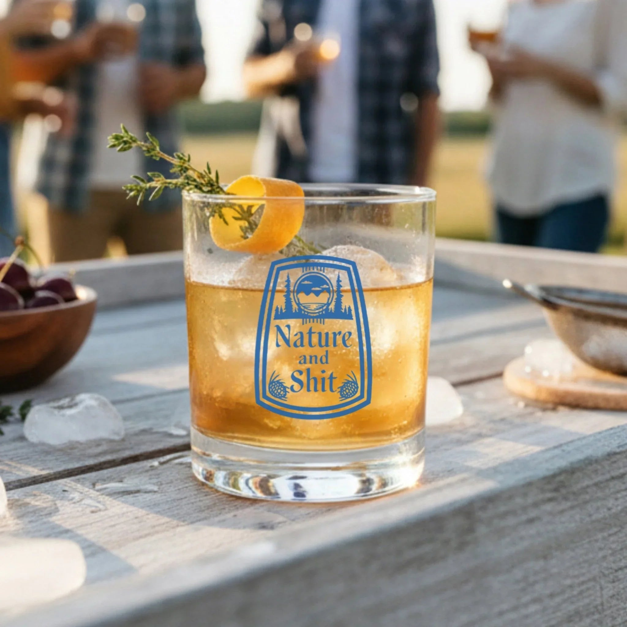 A glass of amber-colored liquid, possibly a cocktail, with a slice of lemon and a sprig of rosemary, sits on a wooden table.