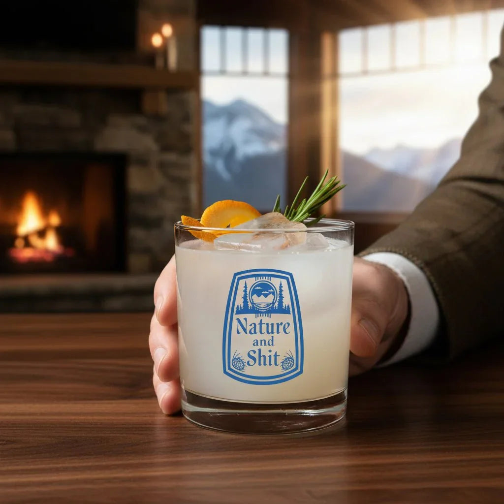 A person's hand holding a glass with a cocktail garnished with an orange slice and a sprig of rosemary, with a fireplace and mountains visible in the background.