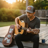 A man is sitting on a wooden deck, playing a guitar and holding a cup of coffee.