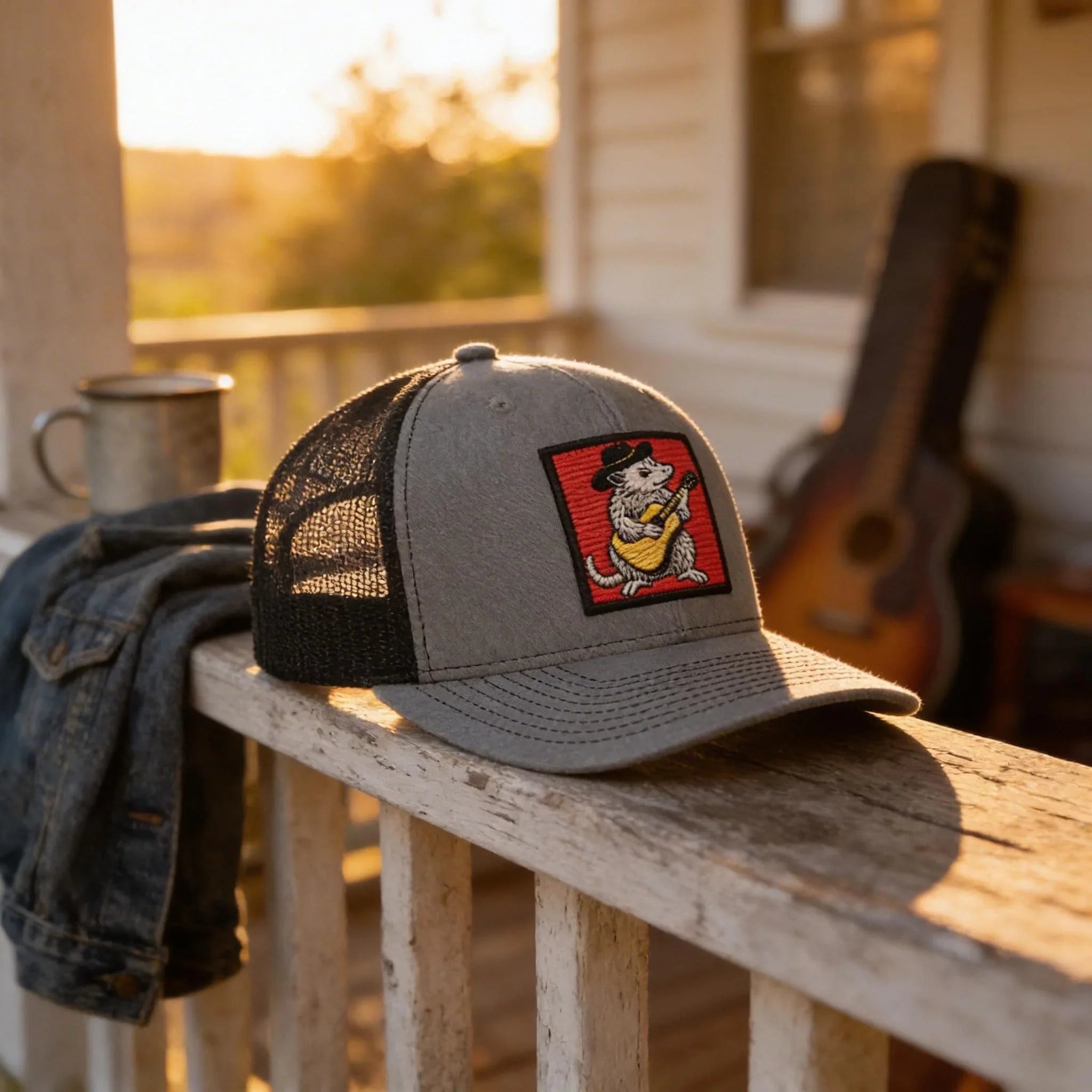 a gray baseball cap with a red and black patch on the front, placed on a wooden railing. In the background, there is a guitar and a mug, suggesting a relaxed outdoor setting.