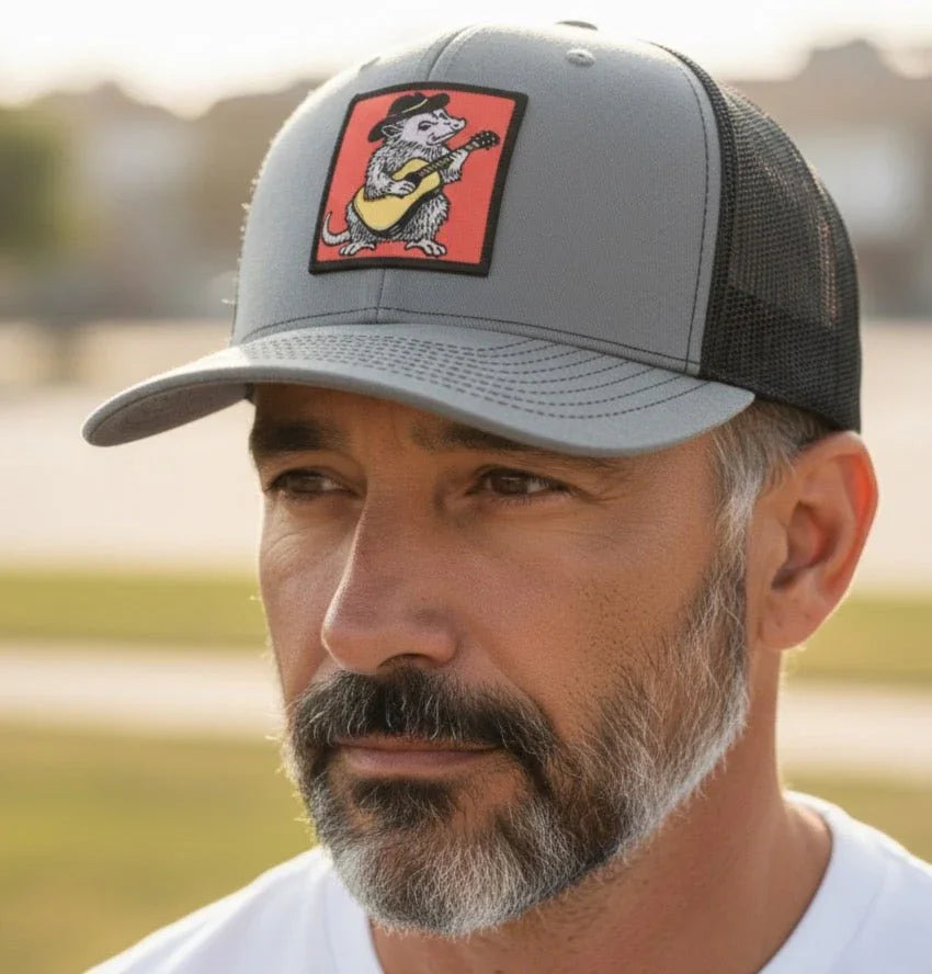A man wearing a gray baseball cap with a cartoon image of a cat playing a guitar on it.