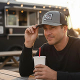 A man wearing a gray baseball cap with a logo is sitting at a table, holding a white cup with a red straw.