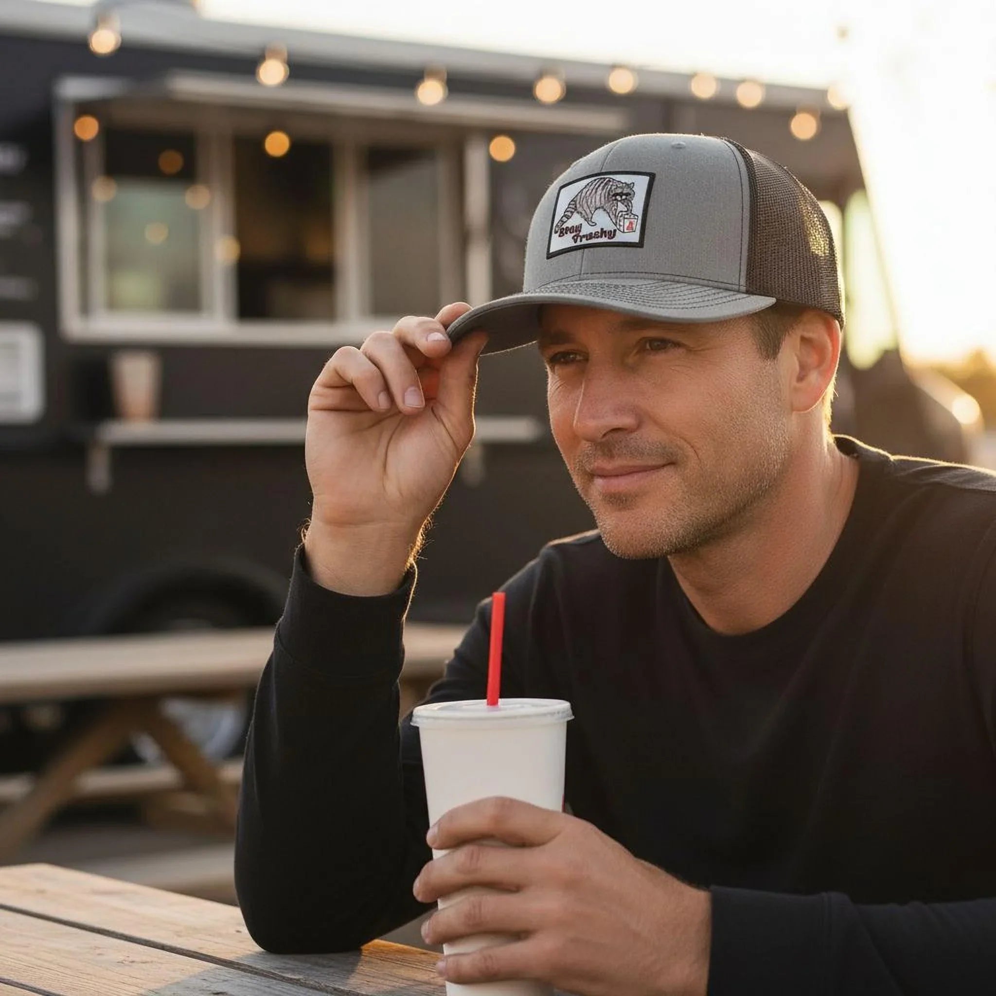 A man wearing a gray baseball cap with a logo is sitting at a table, holding a white cup with a red straw.