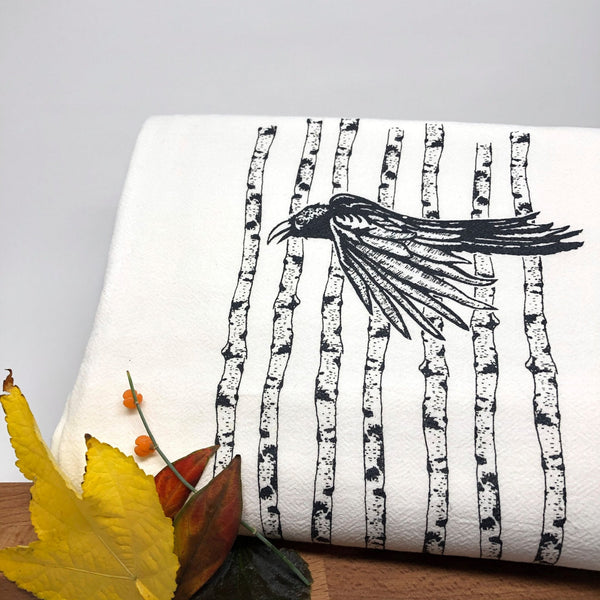 Black raven tea towel artistically arranged with autumn leaves in the background, emphasizing the beautiful aspen tree design.
