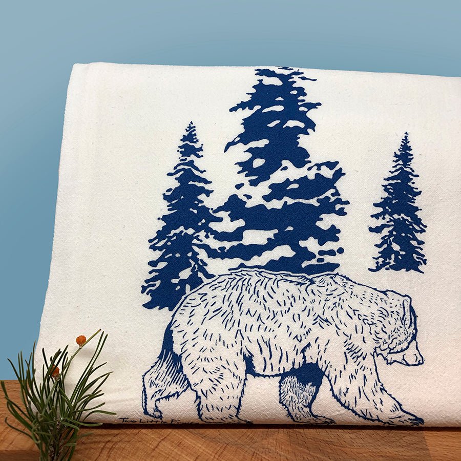 Bear kitchen towel featuring a blue bear design walking through pine trees, elegantly folded on a wooden surface.