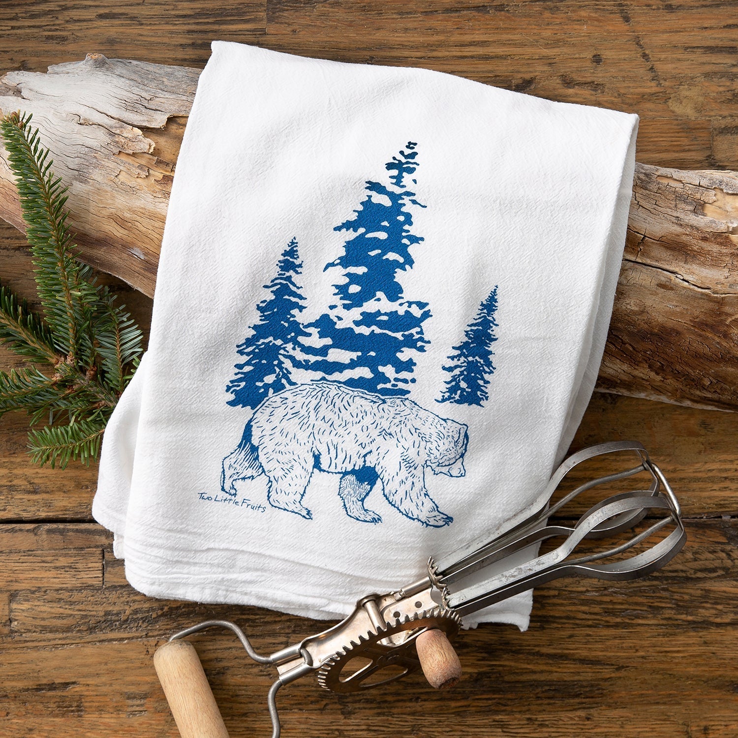 Bear kitchen towel with a blue bear image and pine trees, placed on a rustic wooden background with kitchen utensils.