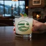 A hand is holding a glass containing a cocktail garnished with a lime and a sprig of mint.