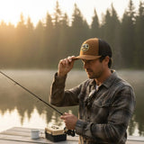 A man in a plaid shirt and baseball cap stands on a dock, holding a fishing rod and looking at his reflection in the water.