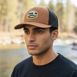 A man wearing a brown baseball cap with a patch on it, standing in front of a body of water.