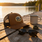 A brown baseball cap with a fish patch, sunglasses, and a mug are arranged on a wooden dock overlooking a lake at sunset.
