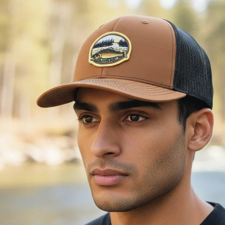 Man wearing a brown and black cap with a logo, standing outdoors near water and trees.