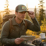 A man in a green jacket and baseball cap sits on a rock, holding a map and looking at it while wearing a backpack.