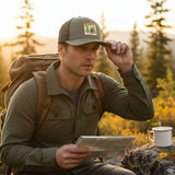 A man in a green jacket and baseball cap sits on a rock, holding a map and looking at it while wearing a backpack.