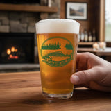 A hand is holding a glass of beer with a green design featuring a fish and a landscape, with a fireplace visible in the background.
