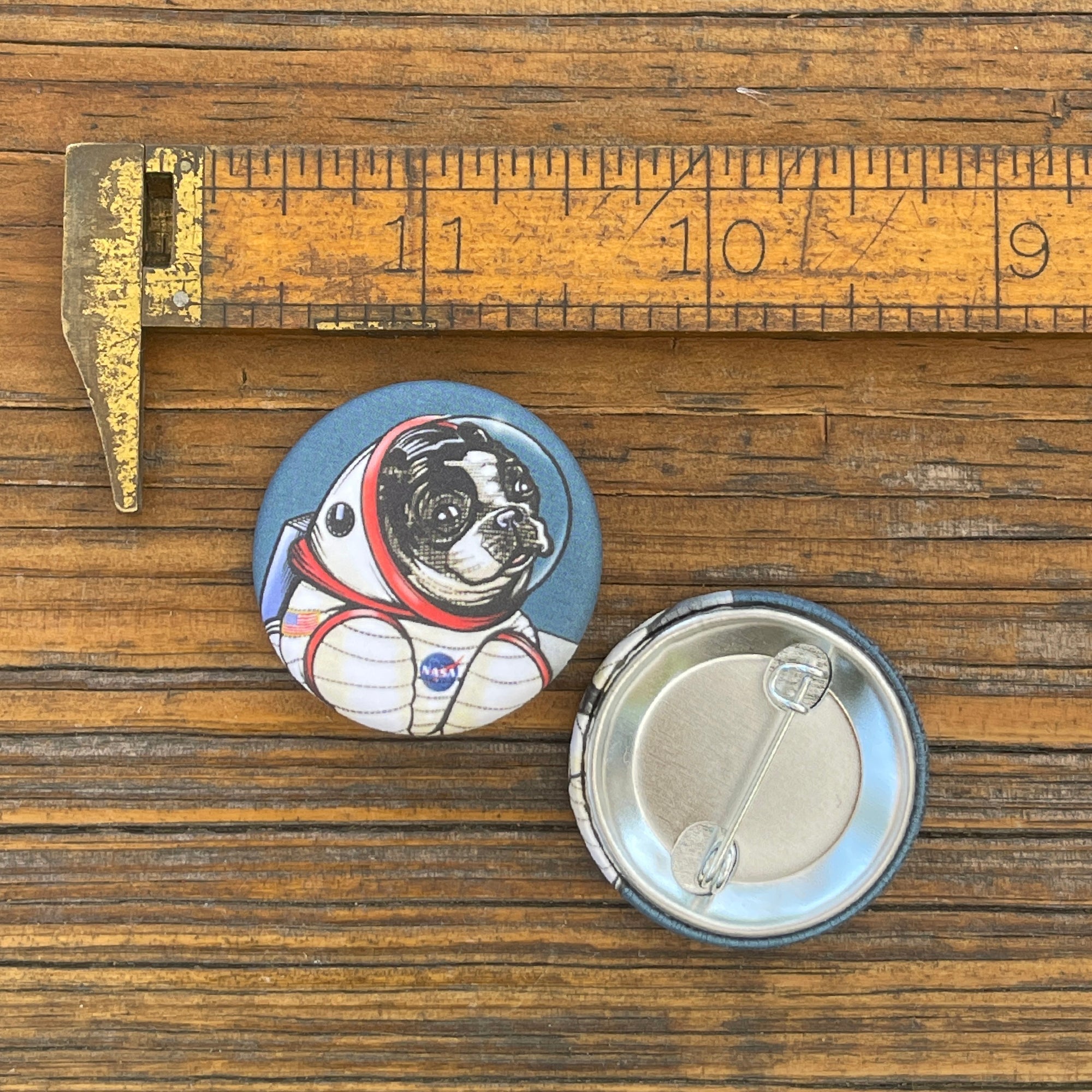 Astronaut dog button pin shown next to a ruler, highlighting its 1.25 inch size; the design features a dog in a space suit with a NASA logo.
