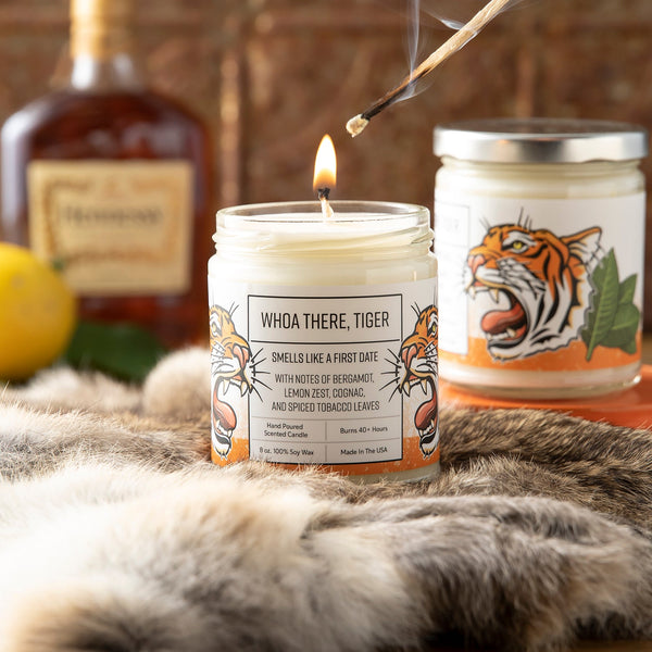 A hand lighting the wick of the 'Whoa There, Tiger' candle with bergamot lemon fragrance, showcasing its enticing scent blend, surrounded by a backdrop of lemon and cognac.