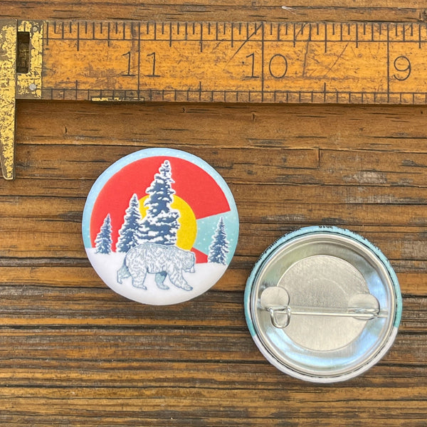Colorado flag pin featuring a bear design, measuring 1.25 inches, displayed next to a ruler on a wooden surface.