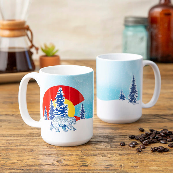 Two Colorado bear mugs on a wooden table, displaying a snowy mountain scene with a bear, perfect for coffee enthusiasts and nature lovers.