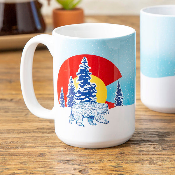 Close-up view of a Colorado bear mug showcasing a vibrant bear design with snowy trees and a colorful sunset, emphasizing its unique style and 15 oz capacity.