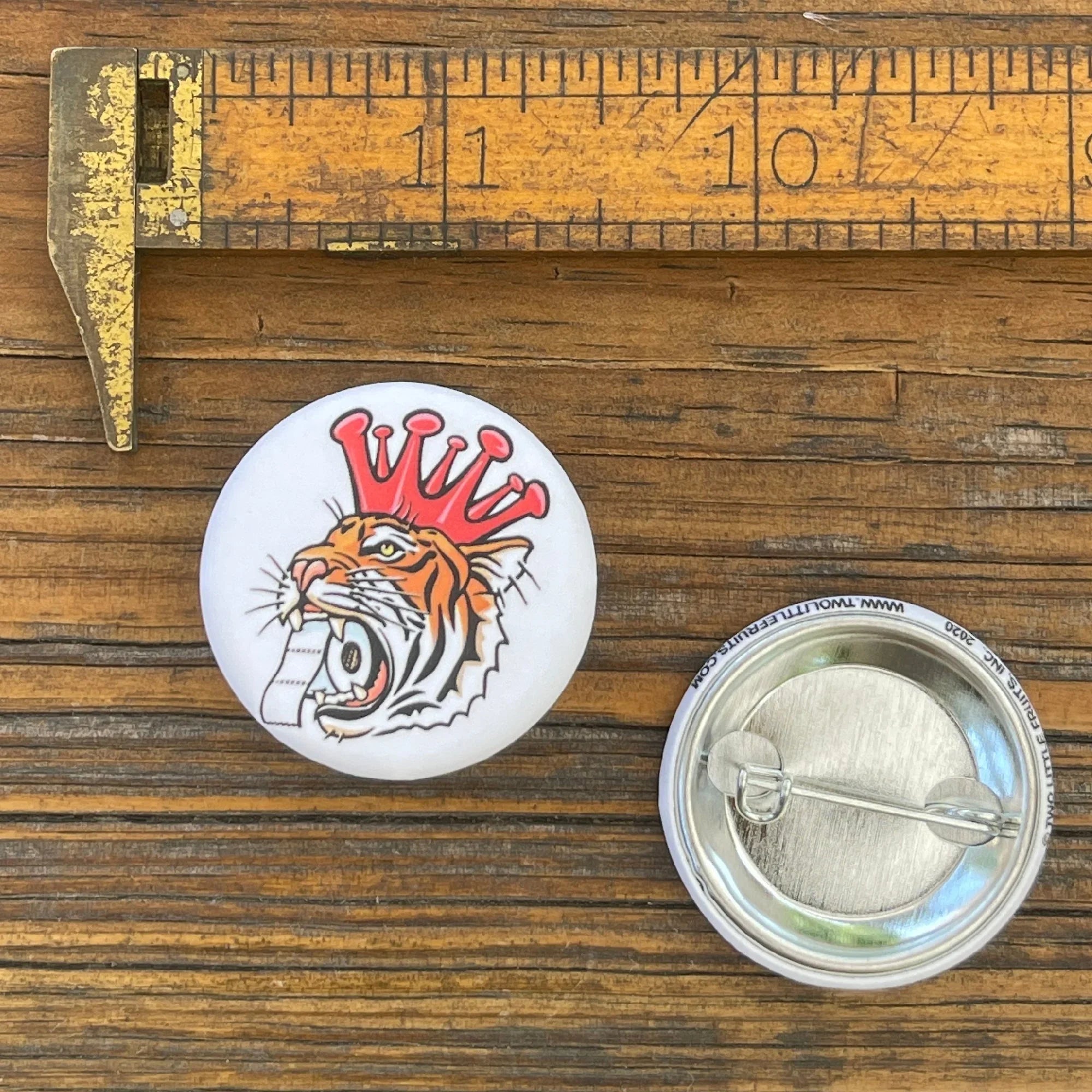 Corona King Tiger Button Pin measuring 1.25 inches, showcasing a vibrant tiger design with a crown, resting next to a ruler on a wooden surface.