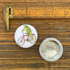 Frog Button Pin - Toadster The Frog, measuring 1.25 inches, showcasing a playful illustration of a frog riding a bike next to a ruler on a wooden background.