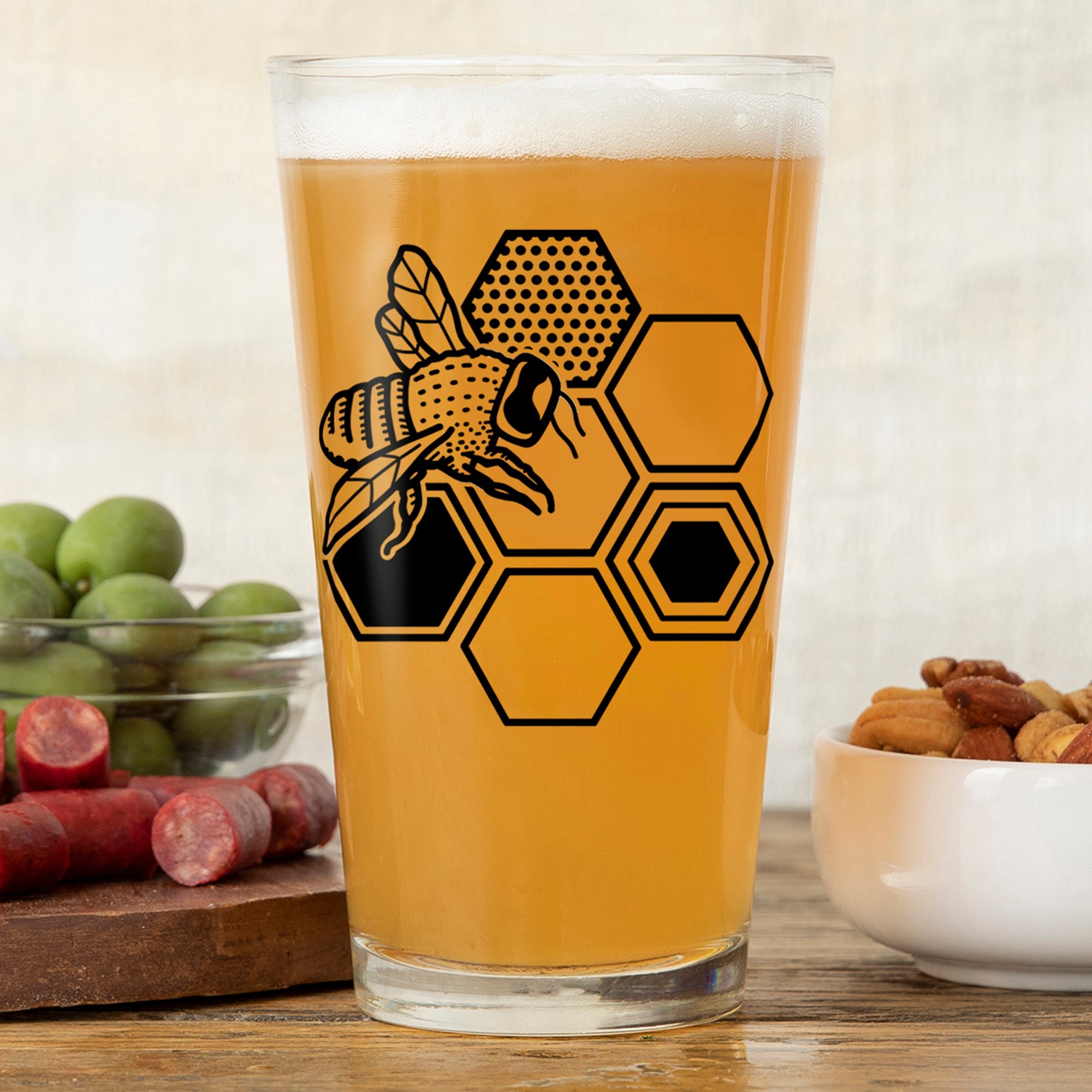 Enjoy your drinks in this stylish bee beer glass, featuring a vibrant bee and honeycomb design with a background of snacks.