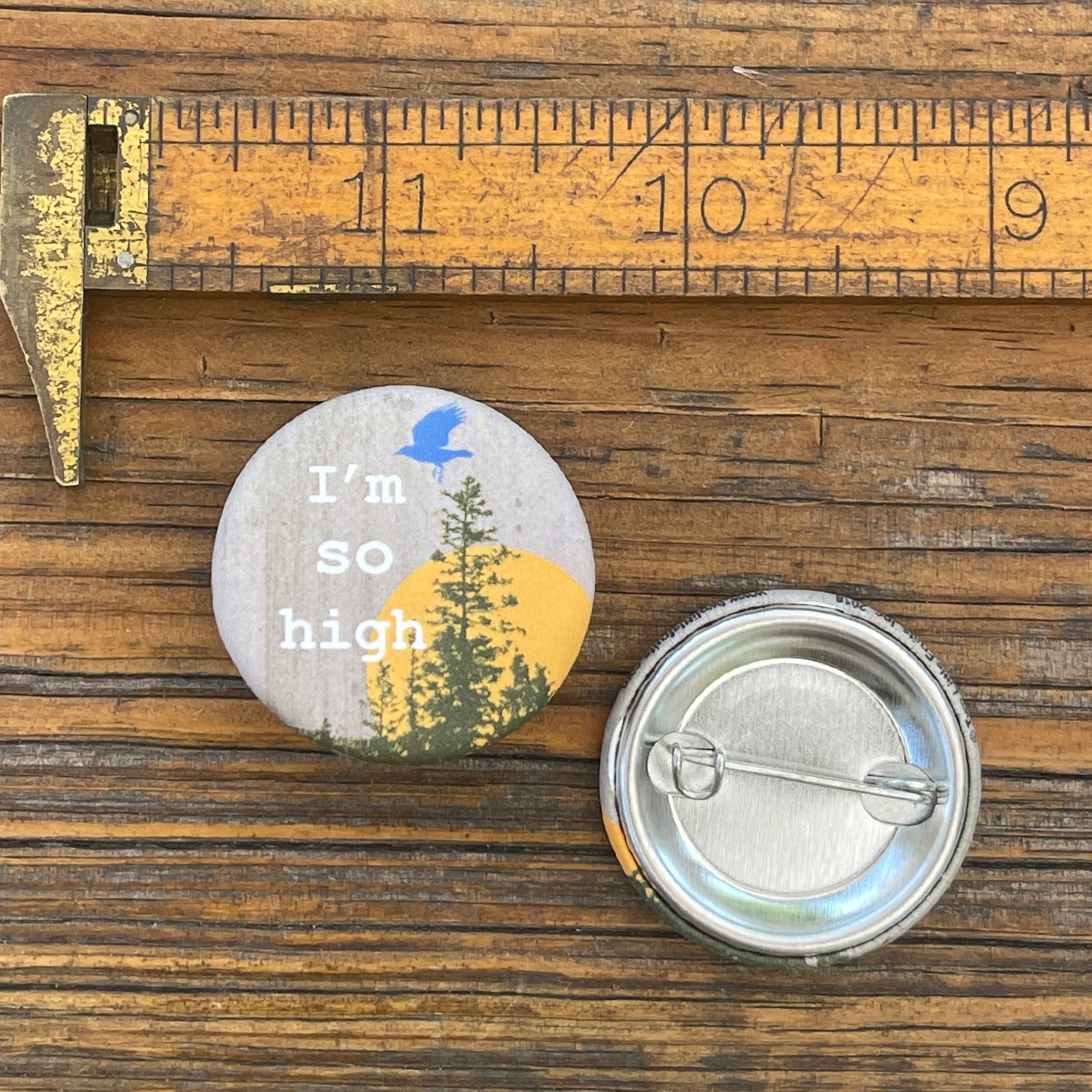 Close-up of a funny button pin reading 'I'm so high' with nature graphics, featuring a bird and tree silhouette, alongside a measurement ruler.