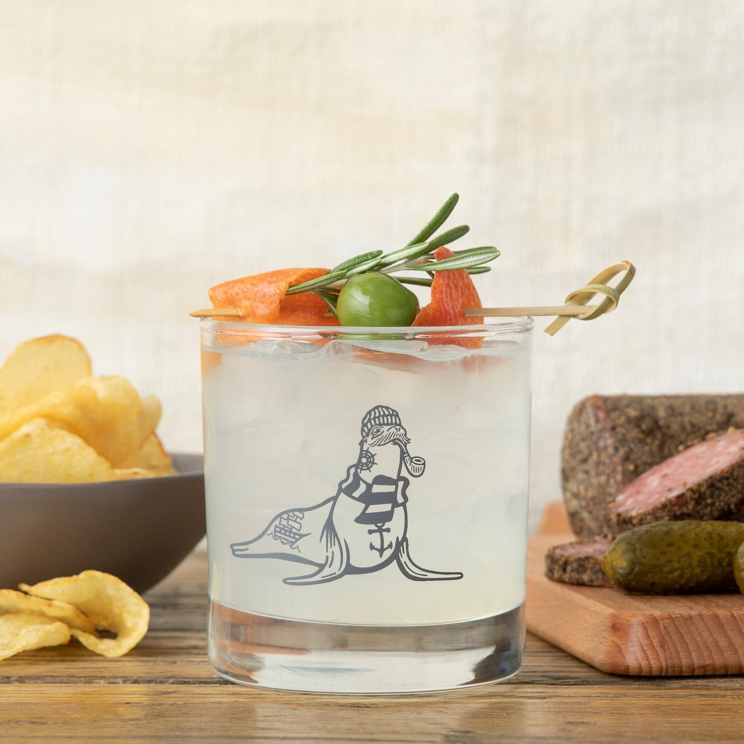 Savor a delightful drink in a whiskey glass from the Mix and Match Your Own 6 Unique Whiskey Glass Set, featuring an elegant sea lion illustration, complemented by tasty snacks.
