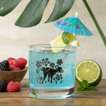 A vibrant blue cocktail in a whiskey glass from the Mix and Match Your Own 6 Unique Whiskey Glass Set, adorned with a playful design of flowers and a charming dog.