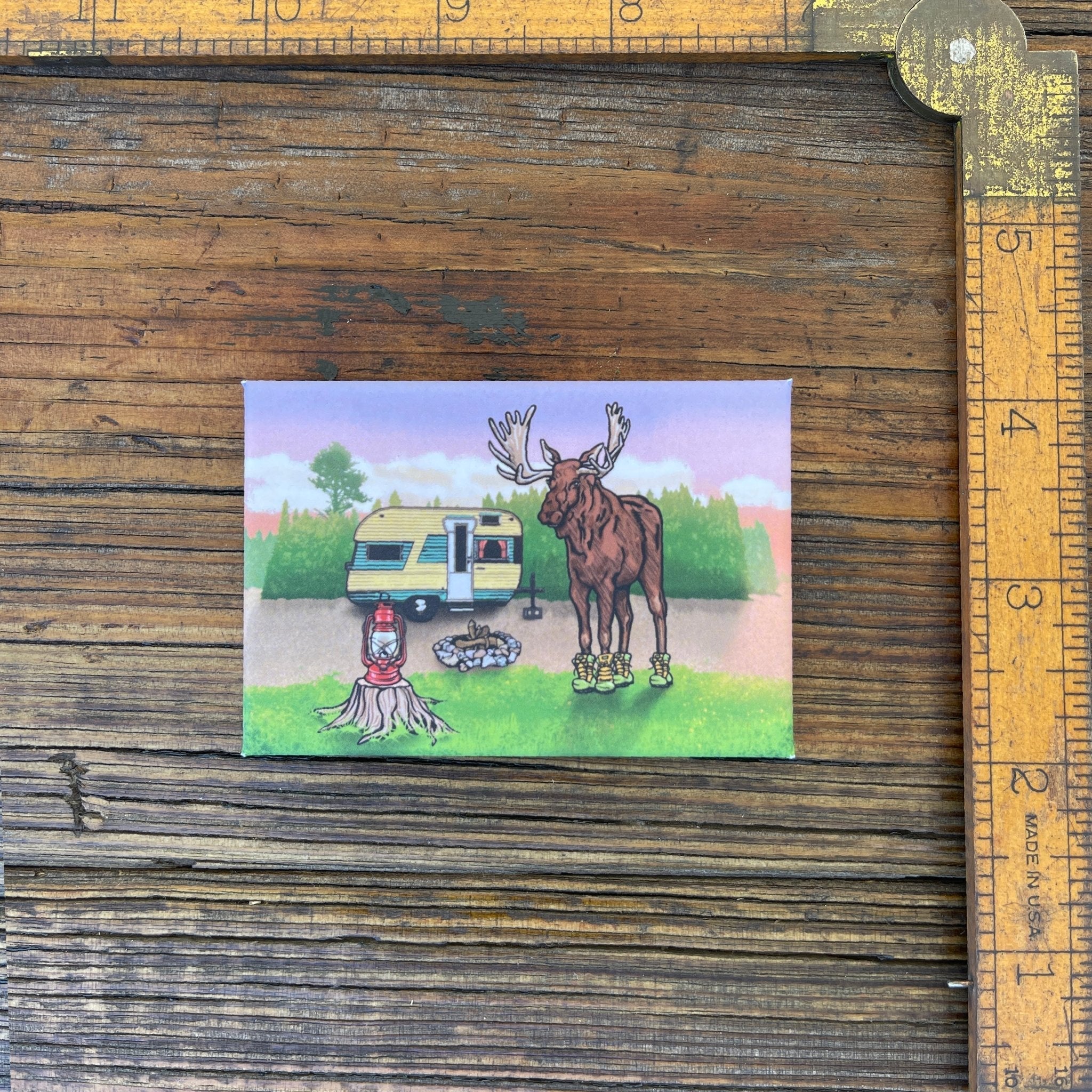 Moose In Boots Fridge Magnet next to a ruler, showcasing its charming design with a moose by a camper, ideal for moose themed decor.