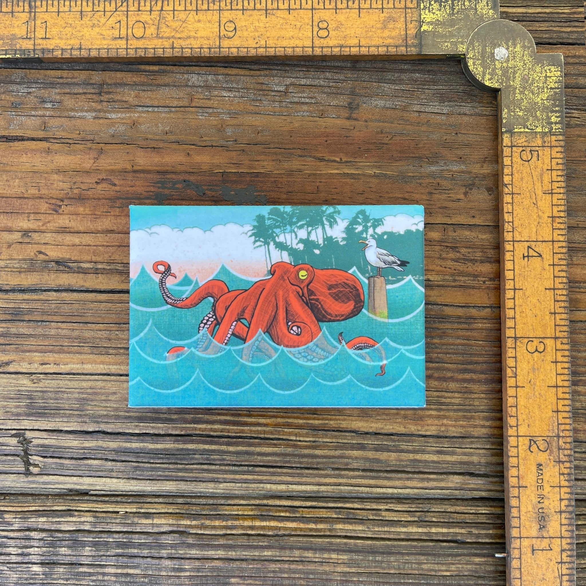 Charming octopus fridge magnet illustrated with a red octopus surrounded by ocean waves, measuring 2" x 3" for a delightful kitchen accessory.