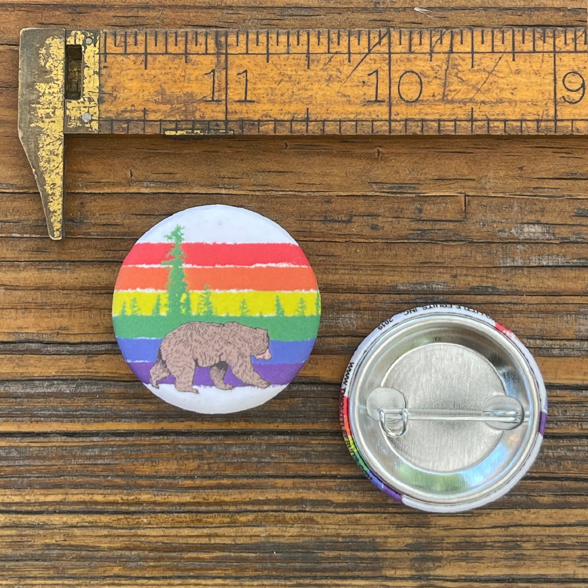 A rainbow bear pin displayed next to a measuring scale for size reference, showcasing its vibrant design with a bear set against colorful stripes.