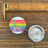 A rainbow bear pin displayed next to a measuring scale for size reference, showcasing its vibrant design with a bear set against colorful stripes.