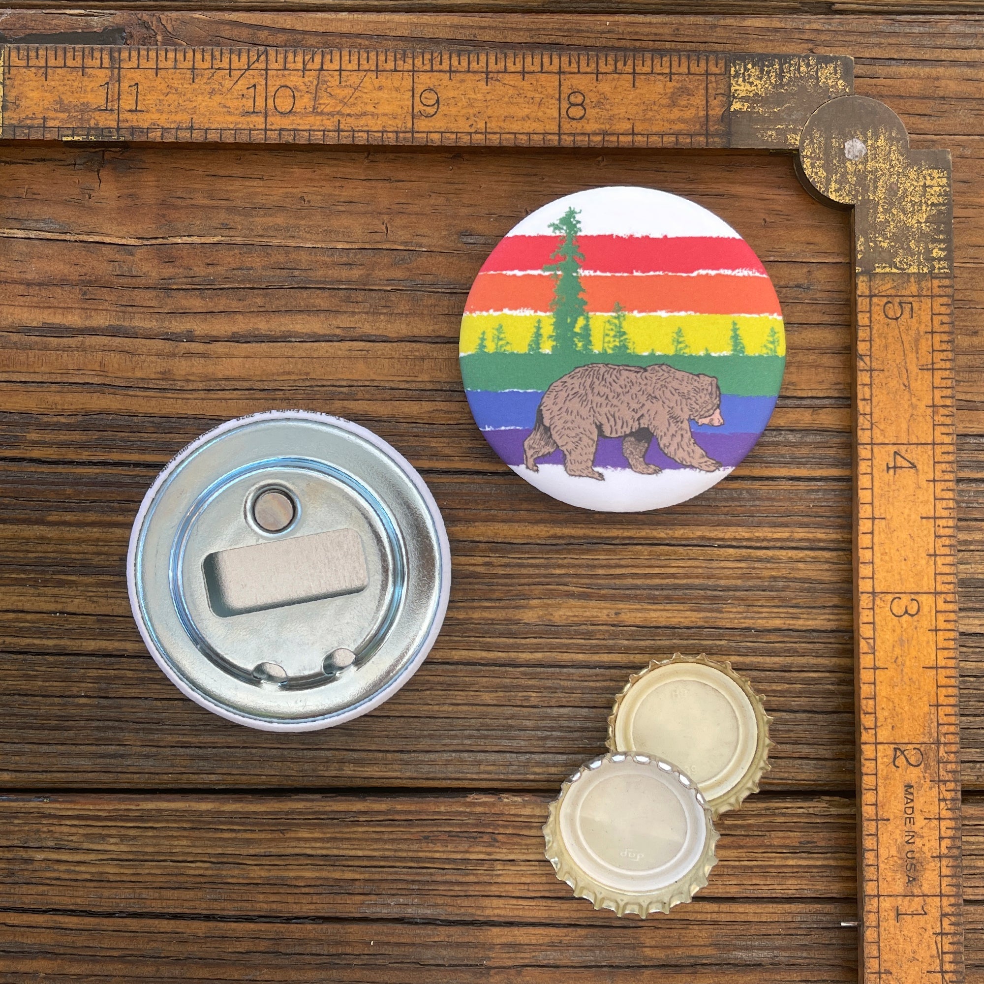 A gay pride bottle opener featuring a colorful bear design against a rainbow background, placed on a wooden table with a ruler and bottle caps.