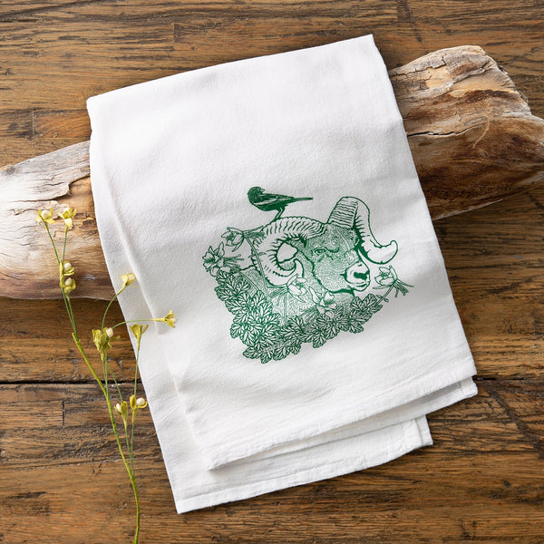 Bighorn Ram Tea Towel displayed on a rustic wooden surface, showcasing its beautiful design of a bighorn ram surrounded by flowers and a bird.
