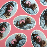 Colorful Sea Otter Stickers arranged on a pink background, showcasing the playful otter with a doughnut.
