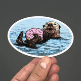 Hand-held Sea Otter Sticker depicting an adorable otter holding a pink sprinkle doughnut in water.
