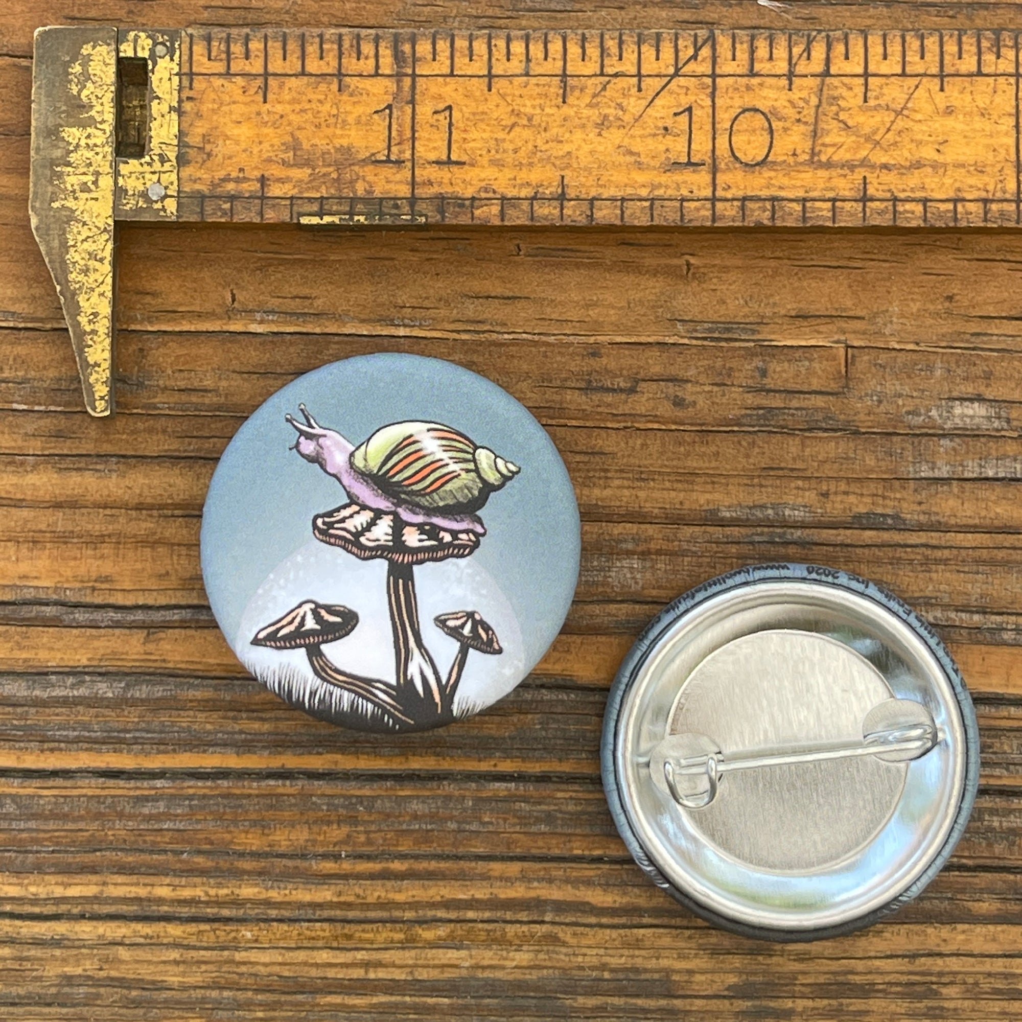 Snail button pin displayed next to a measuring tool on a wooden surface, showcasing its size and design featuring a snail on a mushroom.