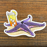 Tipsy Starfish Sticker featuring a purple starfish relaxing on the beach with a refreshing cocktail. Perfect for adding flair to any surface.